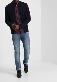 Marinblå stickad cardigan över en röd och blå rutig skjorta, ihop med ljusblå jeans och svarta sneakers. Minimal logotyp på cardigans.