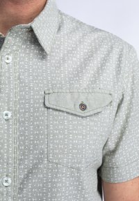 Camisa de manga corta en tejido verde claro con un pequeño patrón geométrico, cuello con botones y un bolsillo en el pecho con un detalle de botón.