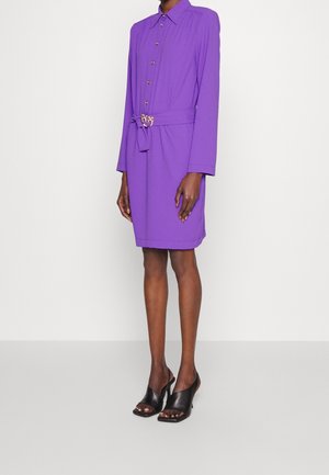 Osoba ubrana w fioletową sukienkę z długim rękawem i guziczkami z przodu, z dopasowanym paskiem oraz czarne klapki na obcasie z odkrytymi palcami, stojąca na jasnym tle.