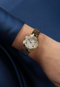 Orologio da polso tono oro con quadrante bianco caratterizzato da numeri romani e piccoli cristalli. Bracciale in metallo regolabile. Cassa rotonda con lunetta testurizzata.