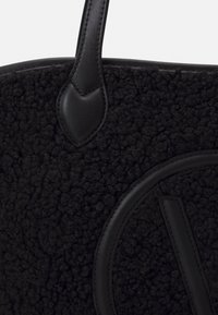 Svart toteväska med en texturerad, fluffig yta och släta läderdetaljer. Har en böjd logotyp och platta läderhandtag.