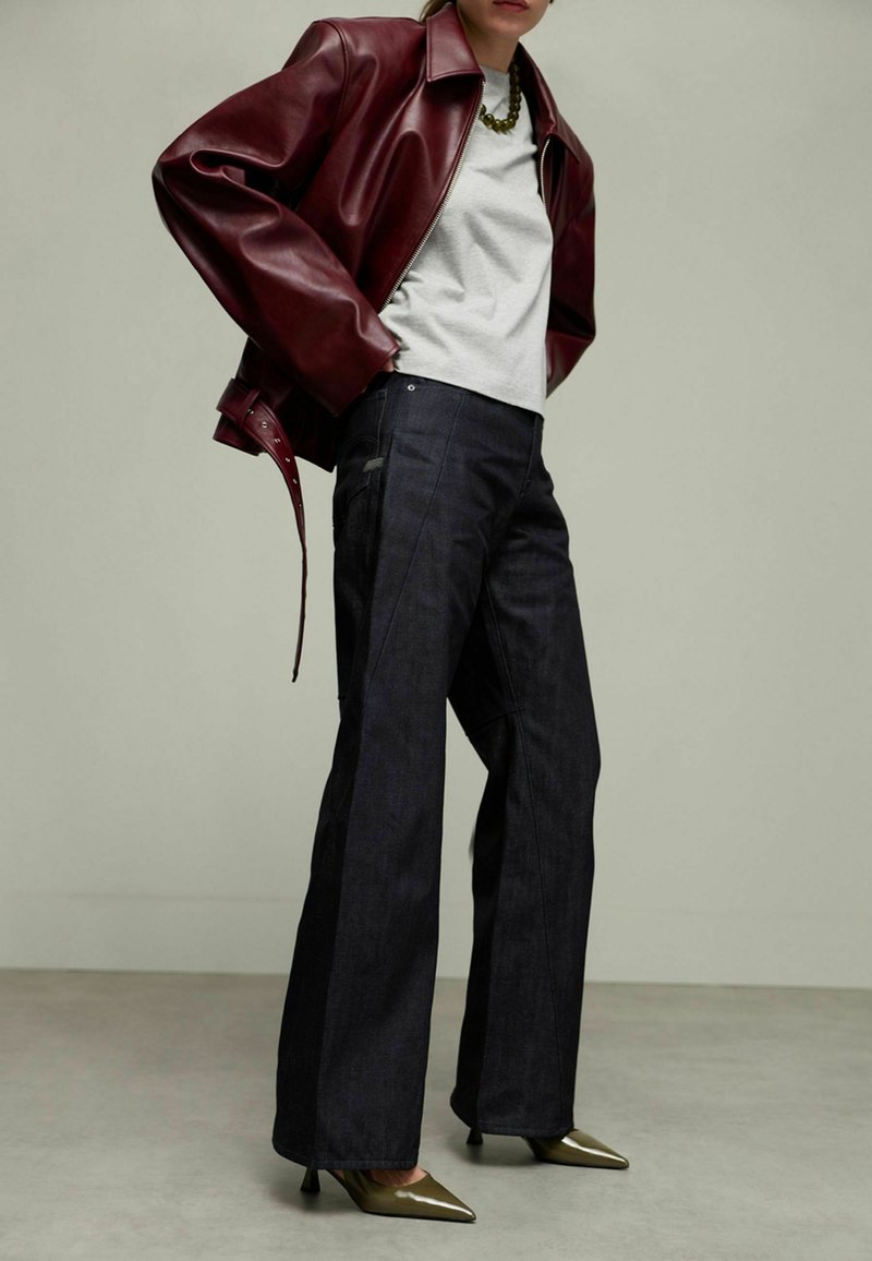 Veste en simili cuir bordeaux, t-shirt gris, pantalons larges en denim foncé, escarpins pointus olive. Détails notables incluant un design de veste superposée.