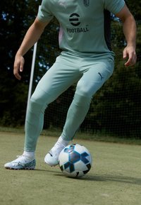 Lichtgroene voetbalkleding met een gestructureerd ontwerp, gecombineerd met witte voetbalschoenen. Een zwart en blauw gestreepte voetbal op een grasveld.