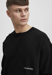 Must värviga dressipluus ümmarguse kaelusega, valmistatud pehmest materjalist. Vasakul küljel on valge tekstdetail. Lihtne, minimalistlik disain.