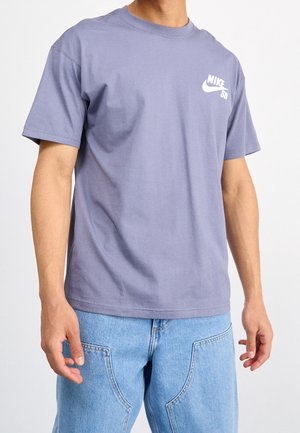 Mand iført en lilla Nike SB t-shirt og lyseblå jeans, vist fra nakke til hofter mod en ensfarvet baggrund.