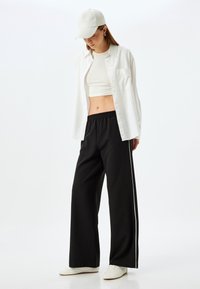 Hvid skjorte med knapper lagdelt over en hvid crop top og sorte bukser med vidde og hvide sidelister. En lysfarvet kasket fuldender outfittet.