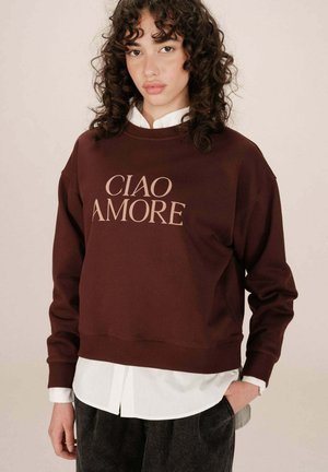 Sweatshirt bordeaux avec "CIAO AMORE" imprimé en beige clair. Dispose d'un col rond, d'épaules tombantes et d'un ourlet côtelé. Porté par-dessus une chemise blanche.