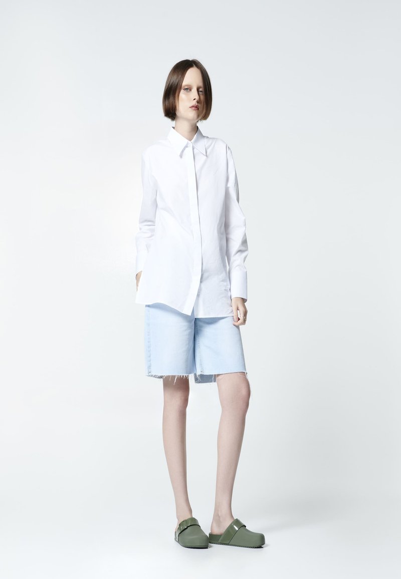 Camisa blanca de botones, pantalones cortos de mezclilla azul claro con dobladillo deshilachado y zapatos verdes sin cordones. Diseño simple con colores lisos y forma ajustada.