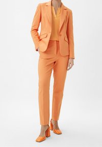 Orange skræddersyet jakkesæt med en tætsiddende blazer og slanke bukser, parret med en lys orange bluse og matchende firkantede hæle.