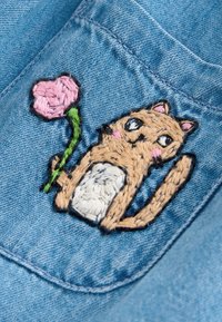 Embroidered tan cat with white belly holding pink flower on blue denim fabric with visible stitching.