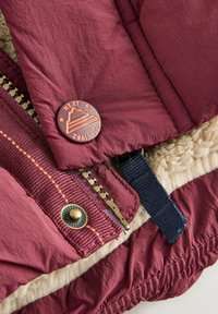 Burgundi pufi dzseki rész, amely szemcsés belső béléssel rendelkezik, krémszínű bolyhos anyaggal, bronz patent gombokkal és arany cipzárral, tengerészkék részletekkel.