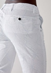 Pantaloni bianchi con ricami floreali blu, design a cinque tasche e un bottone decorativo nero sulla tasca posteriore. Tessuto leggero e strutturato.