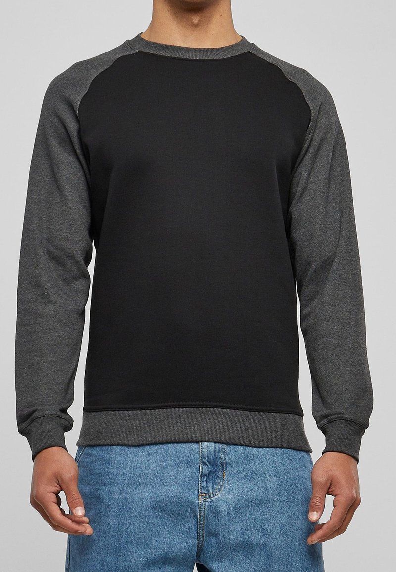 Homme portant un sweat-shirt raglan noir et gris foncé à manches longues, associé à un jean bleu, debout devant un fond clair uni.