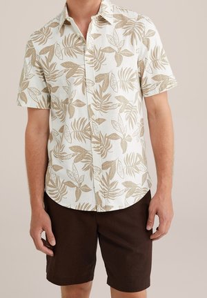 Mężczyzna ubrany w białą koszulę z krótkim rękawem z beżowym liściastym wzorem i ciemnobrązowe szorty, stojący na jednolitym tle.