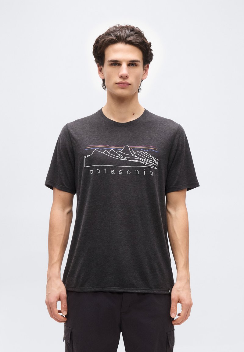 Νέος άντρας που φοράει σκούρο γκρι μπλουζάκι Patagonia με γραμμικό σχέδιο βουνού και έγχρωμες ρίγες, στέκεται μπροστά σε απλό φωτεινό φόντο.