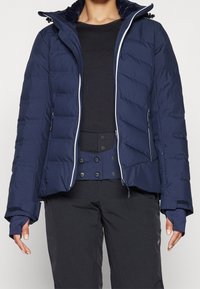 Personne portant une veste matelassée bleu marine avec des poches zippées, un haut noir et un pantalon noir avec un panneau de taille à double bouton.