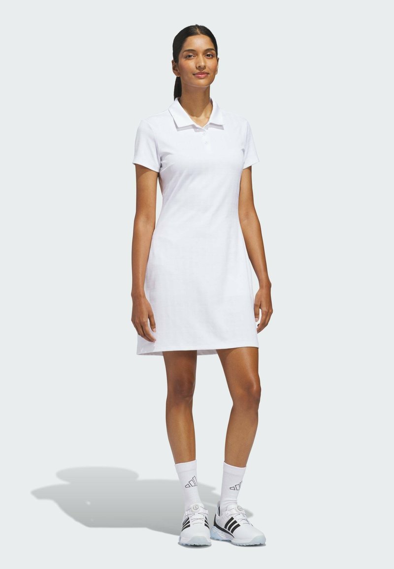 Femme debout portant une robe polo blanche à manches courtes, des chaussettes blanches et des baskets Adidas blanches, sur un fond uni clair.