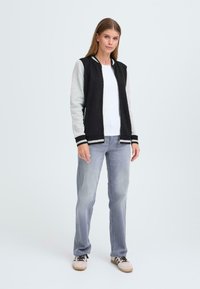 Sort og grå bomberjakke med lynlås foran, ribbede manchetter og strikket krave, båret sammen med lyseblå jeans og beige sneakers.