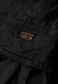 Zwart textiel met een zichtbare structuur en stiksels. Bevat een gestikt etiket met de tekst "SUPERDRY" in gouden en witte letters op een donkere achtergrond.