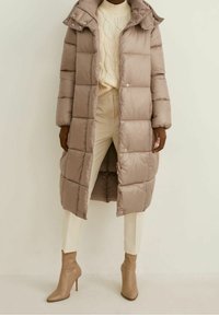 Polstret beige frakke med hætte, med et quiltet design, trykknaplukninger og sidelommer. Båret over en cremede sweater og hvide bukser.