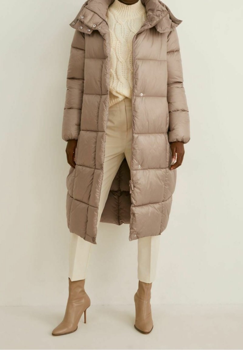 Polstret beige frakke med hætte, med et quiltet design, trykknaplukninger og sidelommer. Båret over en cremede sweater og hvide bukser.