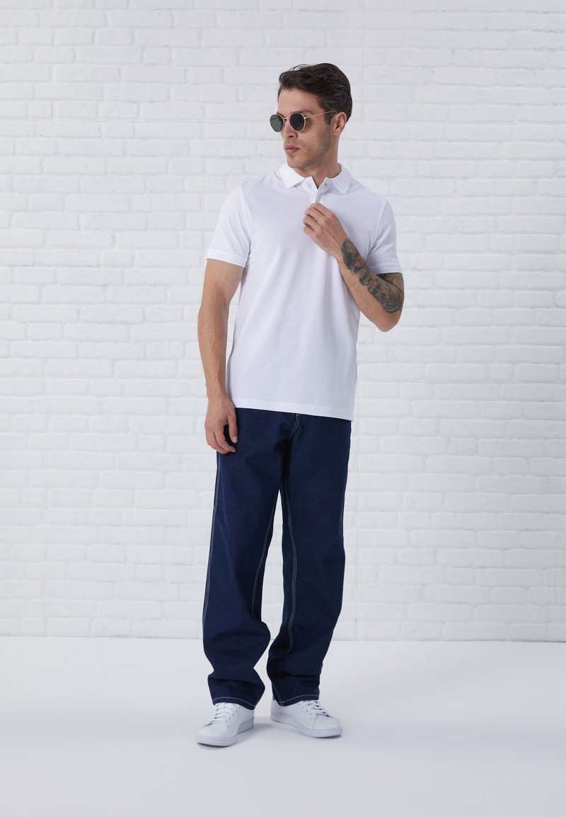 Hvid polo-shirt, mørkeblå brede jeans, hvide sneakers og runde solbriller. Baggrunden har en hvid murstensvæg.