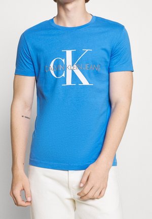 T-shirt bleu à manches courtes en coton avec un col rond. Il présente un logo blanc "CK" et le texte "CALVIN KLEIN JEANS" en gris.