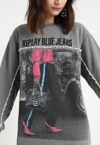 Sudadera gris con un estampado gráfico de una persona en rosa y negro, con detalles en perlas y unos tacones altos de color rosa brillante.