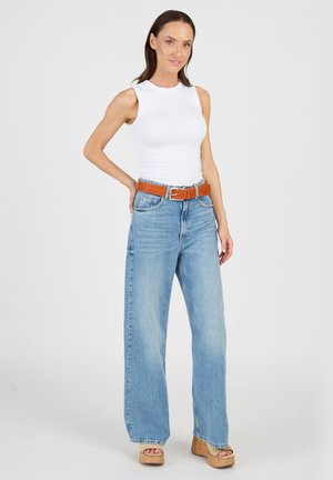 Valkoinen ilman hiuksia oleva toppi yhdistettynä vaaleansinisiin leveälahkeisiin farkkuihin, joita korostaa ruskea vyö ja vaaleanruskeat alustakengät. Sileä kangas, jossa rento istuvuus.