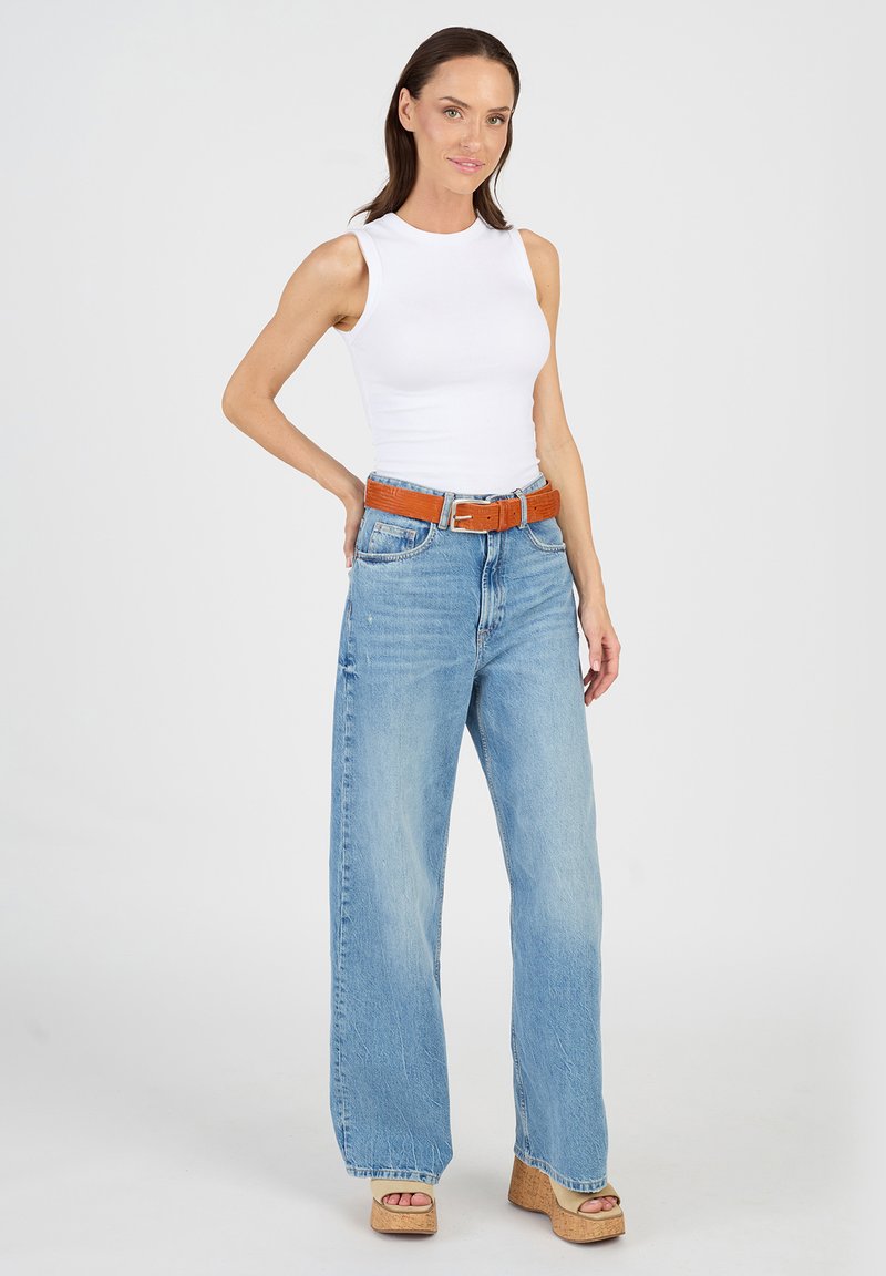 Vit ärmlös topp i kombination med ljusblå vida jeans, accentuerad med ett brunt bälte och klänning med plattformssandaler i beige. Slätt tyg med en avslappnad passform.