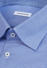 Blå og hvid mønstret skjorte med knaplukket krave og "seidensticker" mærke inde i kraven.