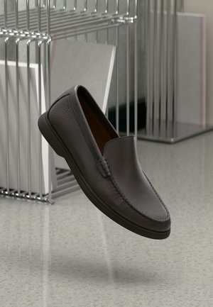 Chaussure en cuir marron foncé à enfiler flottant au-dessus d'un sol poli, avec un présentoir métallique tenant des objets encadrés en arrière-plan.