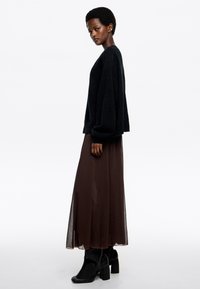 Czarny oversize sweter, zwiewna brązowa przejrzysta spódnica oraz czarne botki na obcasie. Gładka tekstura i nowoczesny, warstwowy design. Widok z profilu.
