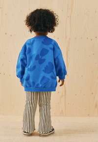 Blaues Sweatshirt mit Blumenmuster, Rippbündchen und elastischem Saum. Kombiniert mit gestreiften Hosen. Hintergrund zeigt eine helle Holzstruktur.