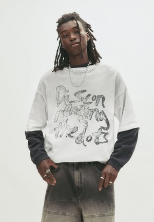 Grå og hvid oversized sweatshirt med sorte ærmer, der har et grafisk tryk med teksten "Discon-necting the dots." Båret over mørke denimshorts.