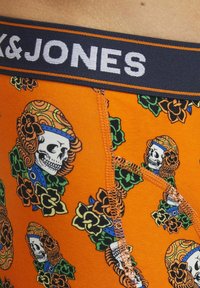 Bóxer naranja con cinta de la marca K&JONES en negro y blanco, que presenta un patrón repetido de calaveras y flores, tejido suave y costuras de contraste.