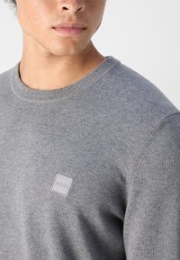 Jeune homme portant un pull gris à col rond avec un petit patch carré "BOSS" sur la poitrine gauche.