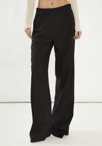 Pantaloni neri su misura con un design a gamba larga, caratterizzati da una parte anteriore piana, pieghe e una consistenza liscia. Realizzati in tessuto leggero.