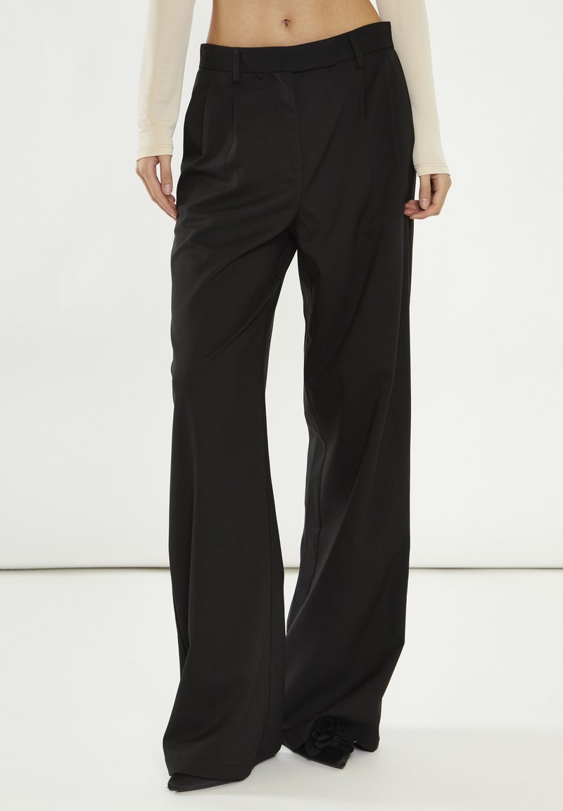 Pantaloni neri su misura con un design a gamba larga, caratterizzati da una parte anteriore piana, pieghe e una consistenza liscia. Realizzati in tessuto leggero.