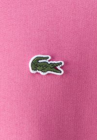 Tissu rose avec une finition texturée présentant un logo crocodile vert brodé, souligné en blanc.