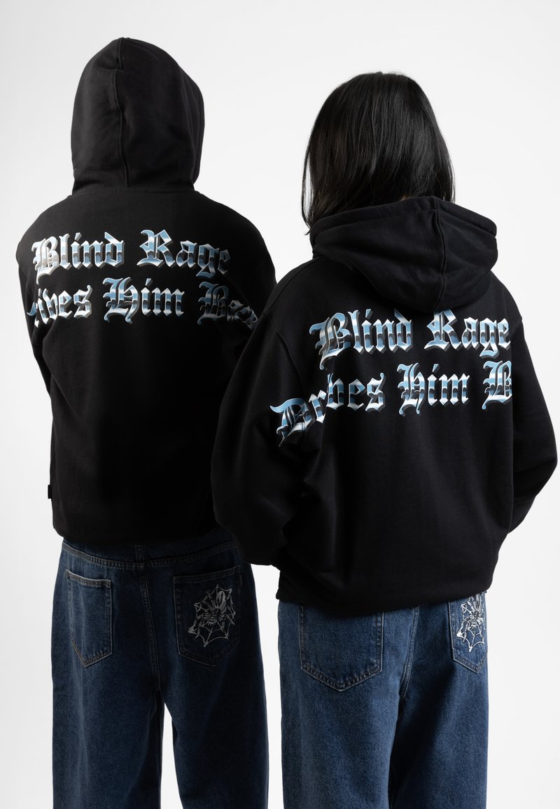 Zwarte hoodie met witte en blauwe tekst op de achterkant, gecombineerd met blauwe jeans met een wolf- en spinnenwebontwerp op de zakken.