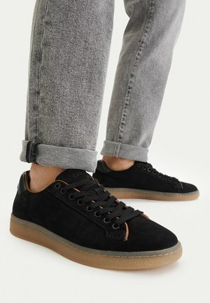 Person trägt schwarze Wildleder-Sneaker mit braunen Sohlen und hochgekrempelte hellgraue Jeans, steht auf einer weißen Fläche.