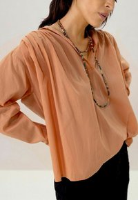 Blouse légère de couleur pêche avec des manches longues, un détail plissé aux épaules et un col en V, accompagnée d'un collier en perles.