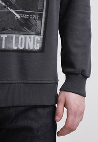 Felpa grigia con polsini e orlo a coste, caratterizzata da una stampa grafica sul davanti, che presenta testo audace e un design di strumento musicale.