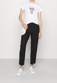 Valkoinen lyhythihainen t-paita graafisella printillä, yhdistetty mustiin muotopestyihin housuihin ja valkoisiin lenkkareihin. Laukku on kermaisen värinen ja sileäpintainen.