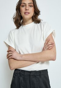 Vit kortärmad topp med hög urringning och vikta manschetter, tillsammans med svarta höga byxor. Slät textur, minimala designdetaljer.