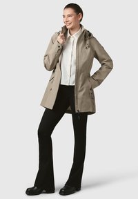 Bežs ūdensnecaurlaidīgs mētelis ar kapuci, ar rāvējslēdzēju priekšā, sānu kabatām un regulējamiem auklām. Apvienots ar melniem plašiem biksēm un melnajām kurpēm.