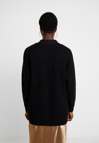 Black knitted sweater with a ribbed texture, long sleeves, and a rounded back hem, worn over a shiny bronze skirt.