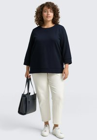 Marineblauer strukturierter Pullover mit Dreiviertelärmeln, kombiniert mit cremefarbenen, schmal zulaufenden Hosen und weißen Sneakers. Hält eine schwarze Tragetasche.