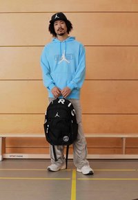 Fekete hátizsák fehér szöveggel és logóval, világoskék kapucnis pulóverrel viselve, amely grafikai mint�ázattal rendelkezik, és szürke melegítőnadrággal.